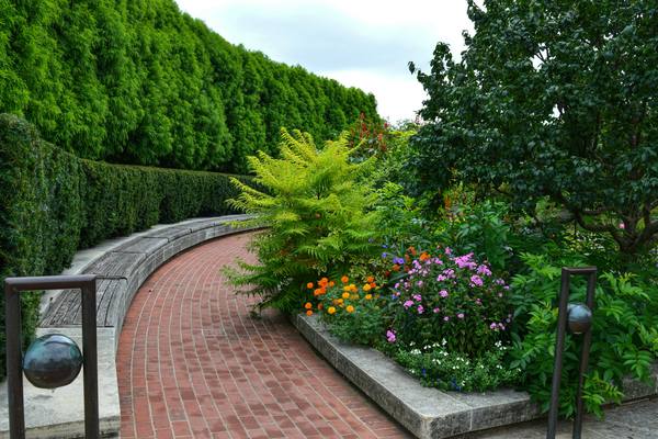 A curved red brick walkway beside a low stone wall, with planted beds of orange and pink flowers against a tall green hedge.