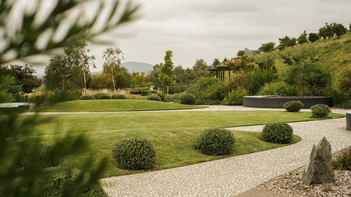 A calm, designed garden: a clipped lawn bordered by curving pale gravel paths, rounded shrubs and layered planting under a soft sky.